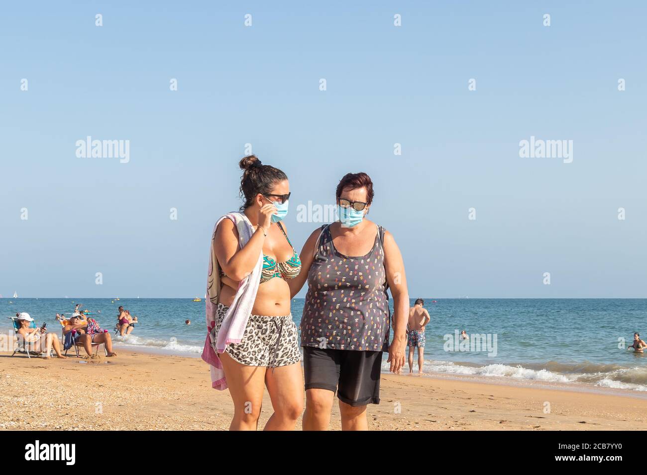 Punta Umbria, Huelva, Spanien - 7. August 2020: Zwei Frauen, die am Strand spazieren gehen und Schutzmasken oder medizinische Gesichtsmasken tragen. Neue Normalität in Spanien mit sozialen Stockfoto