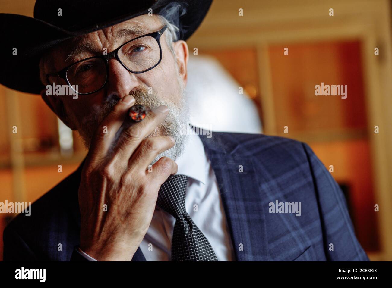 Stilvolle alte - gealterte Dandy in breitrandigen Hut und reichen dunklen Blau mens Suit rauchen Zigarre Indoor, stehend in der Nähe der Theke mit Alkohol Getränke. und l Stockfoto