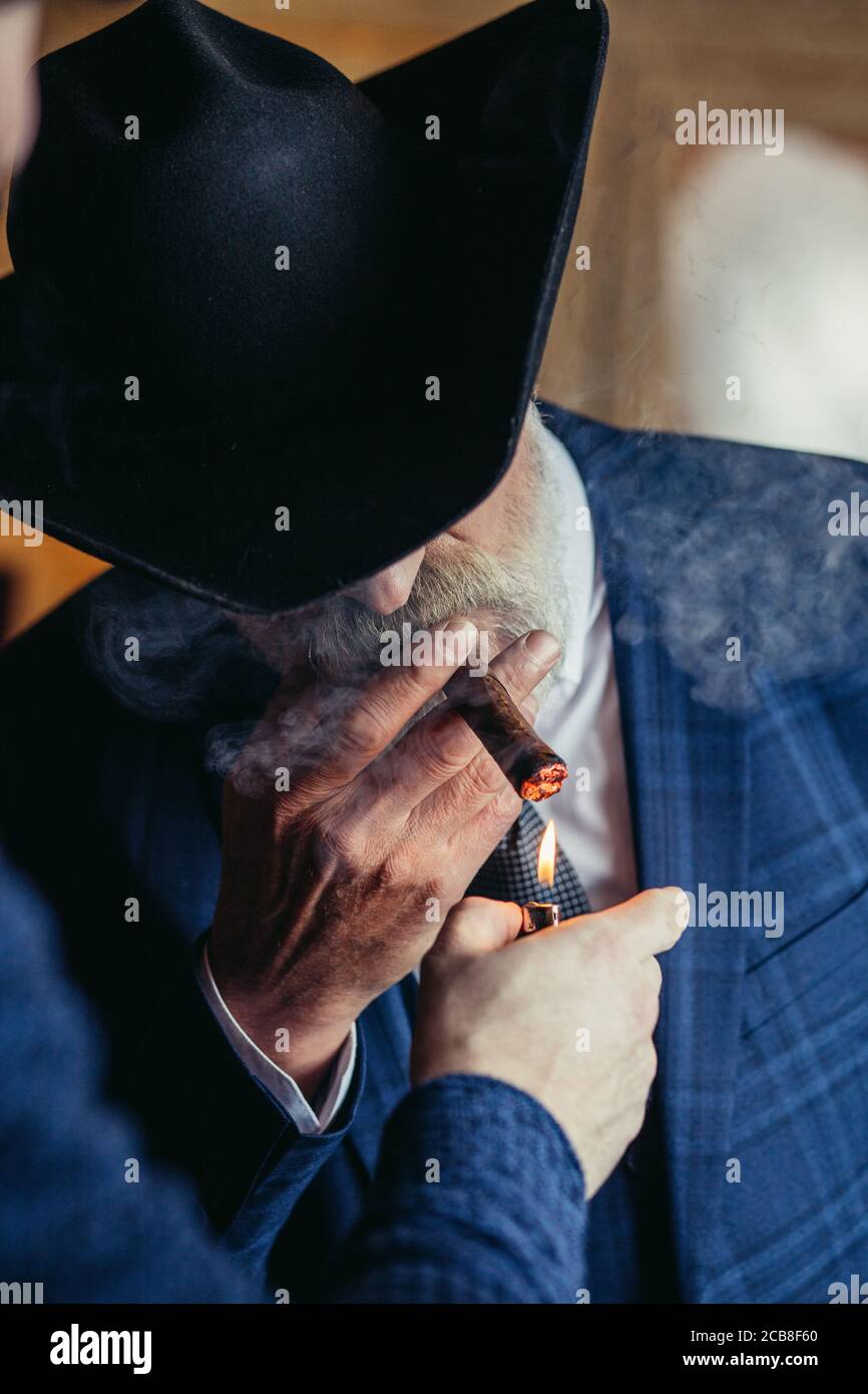 Stilvoller alter Mann mit breitem Krempenhut und dunkelblauem Herrenanzug beleuchtet drinnen eine Zigarre und steht neben der Theke mit alkoholischen Getränken. Stockfoto