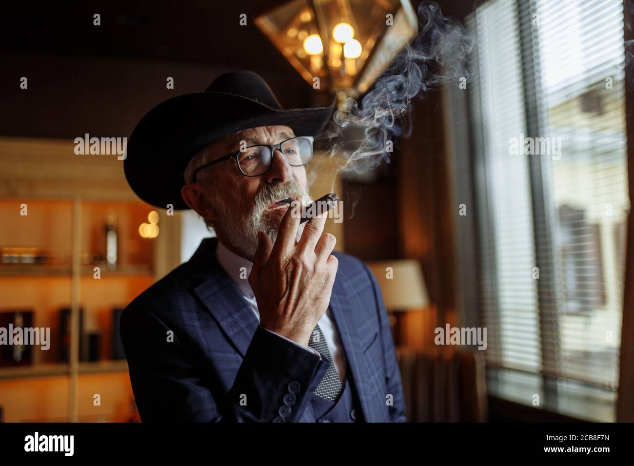 Stilvoller alter Dandy mit breitem Krempenhut und dunkelblauen Herren-Smoking-Zigarren im Innenbereich, der neben der Theke mit alkoholischen Getränken steht. Stockfoto