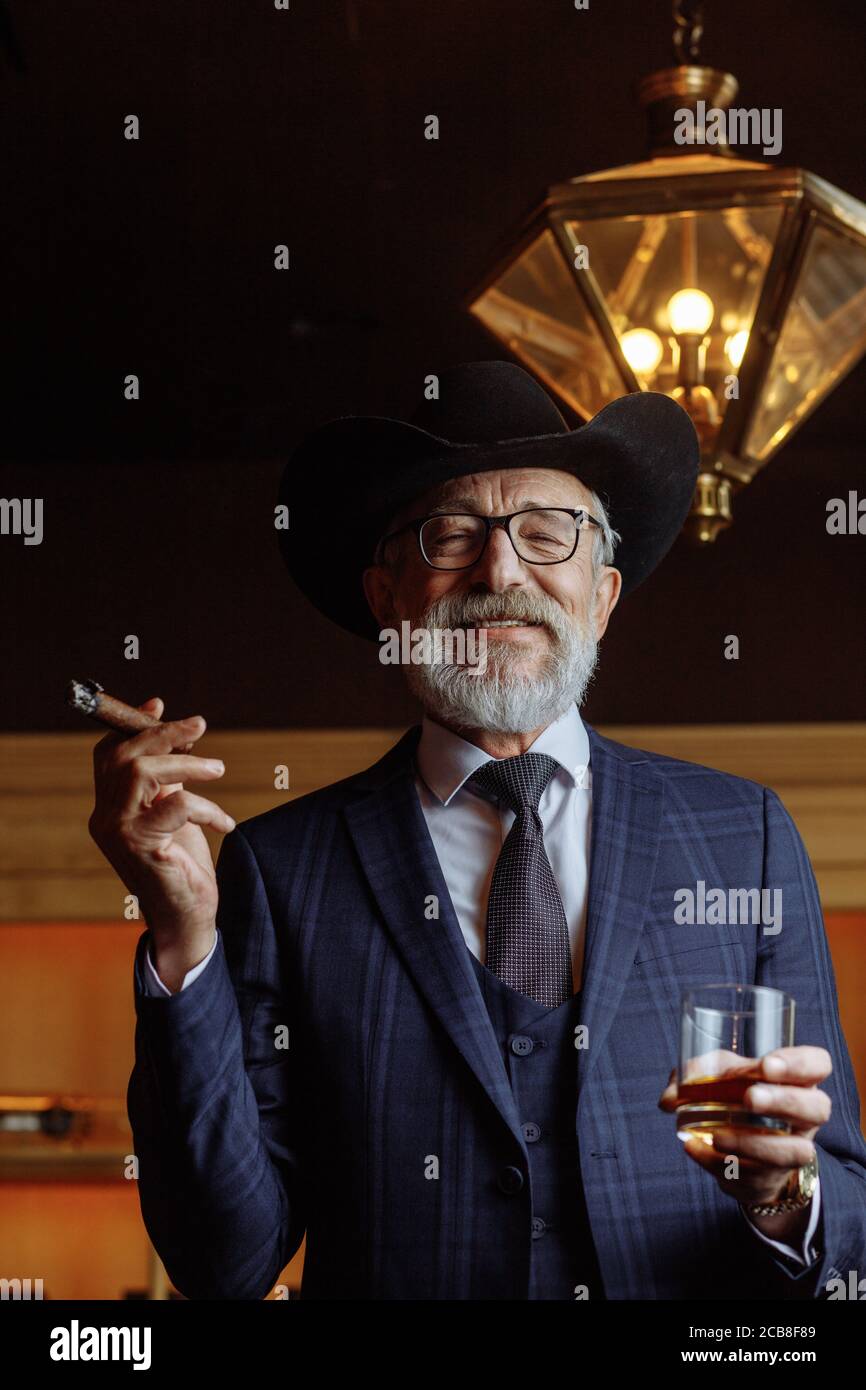 Stilvoller alter Mann in breitem Krempenhut und reichen dunkelblauen Herren Anzug Rauchen Zigarre drinnen, stehen in der Nähe der Theke mit Alkohol Getränke. Und Klo Stockfoto