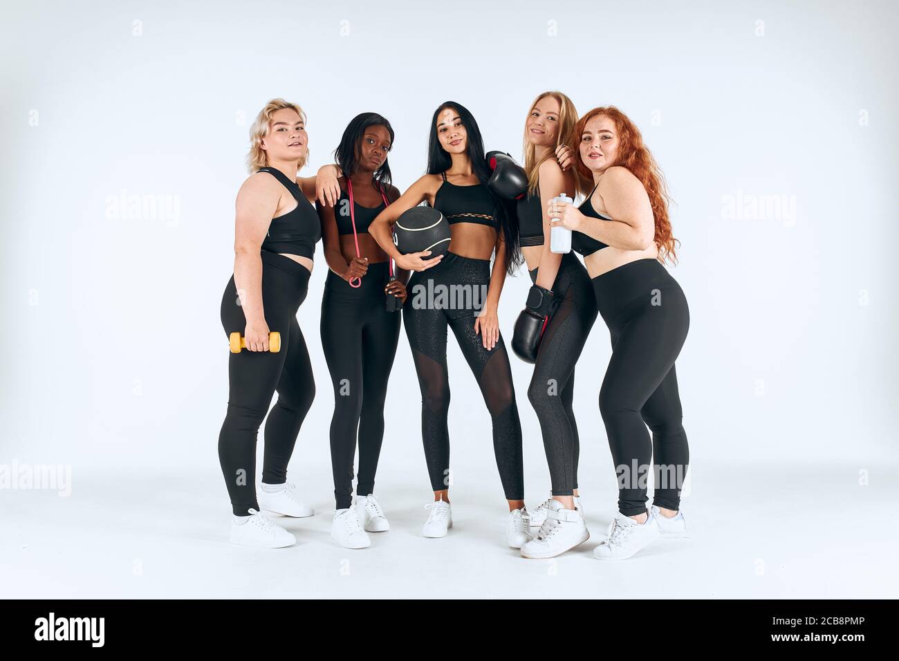 Fünf lächelnde multiethnische Frauen tragen sportliche Leggins und Themen halten Fitness-Ball, Boxhandschuhe und andere Dinge isoliert auf weißem Hintergrund Stockfoto