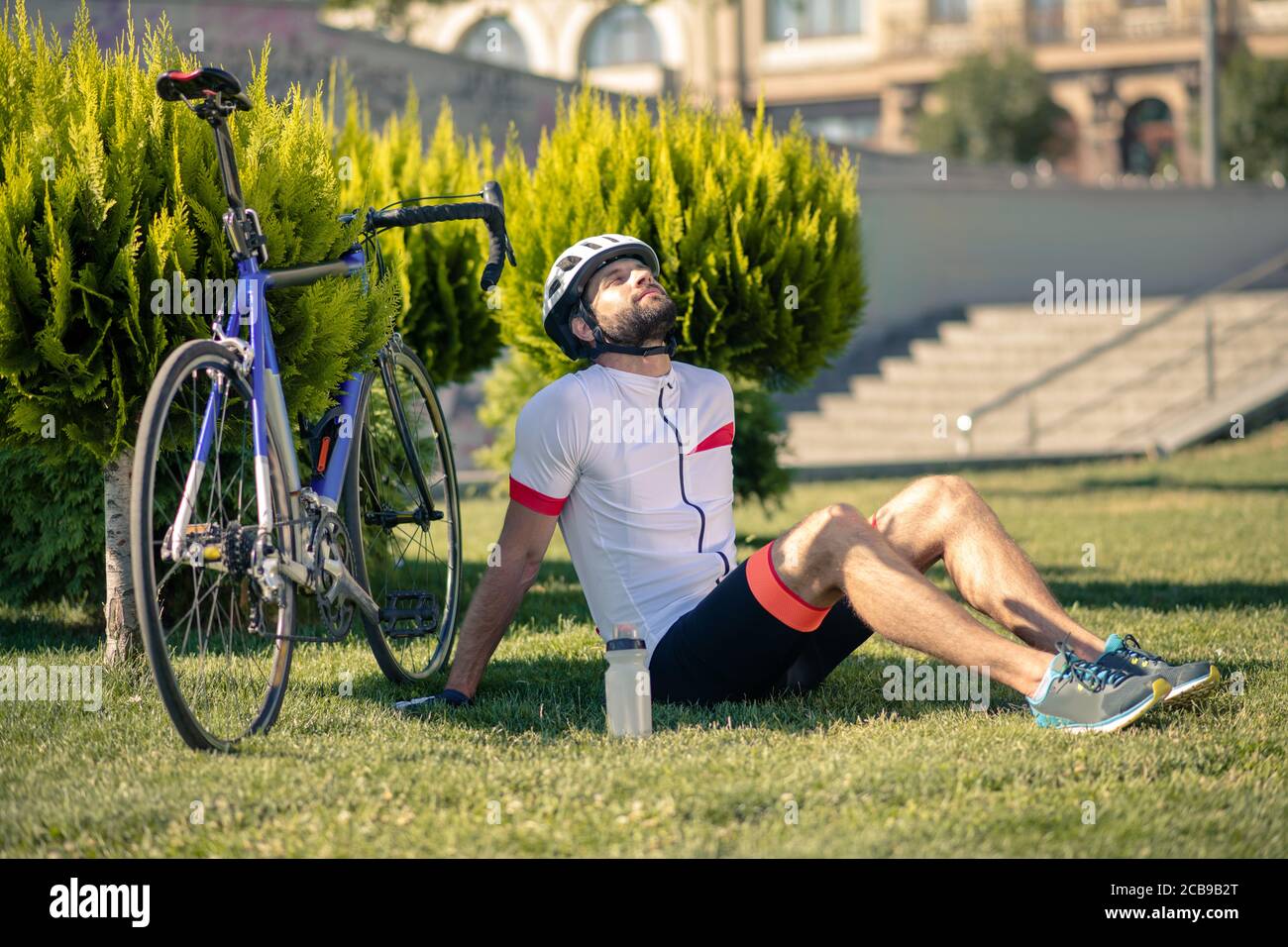 Fröhlicher junger bärtiger Radfahrer sitzt auf Gras Stockfoto