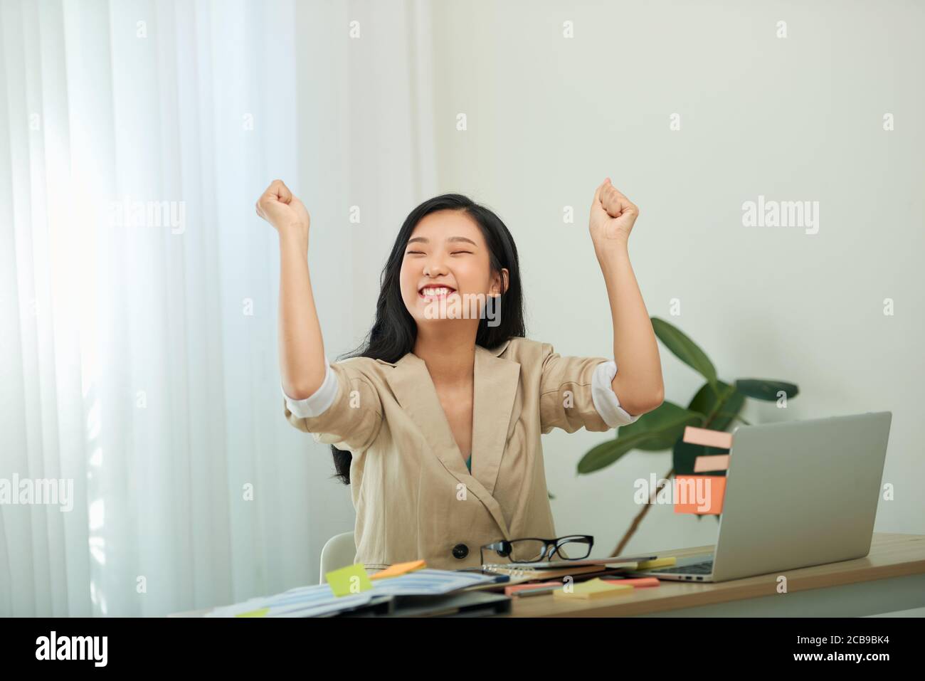 Charmante junge Frau in Brillen sitzen am Tisch in zu Hause Büro mit Laptop, der sich zur Hand lehnt und freudig wegschaut Stockfoto