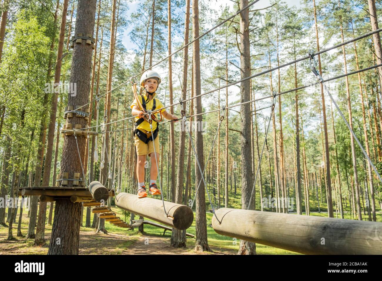 Kleiner Junge mit Kletterausrüstung Kletterseilpfad zwischen Pinien in einem Abenteuerpark. Seilpark. Guten Sommertag. Stockfoto