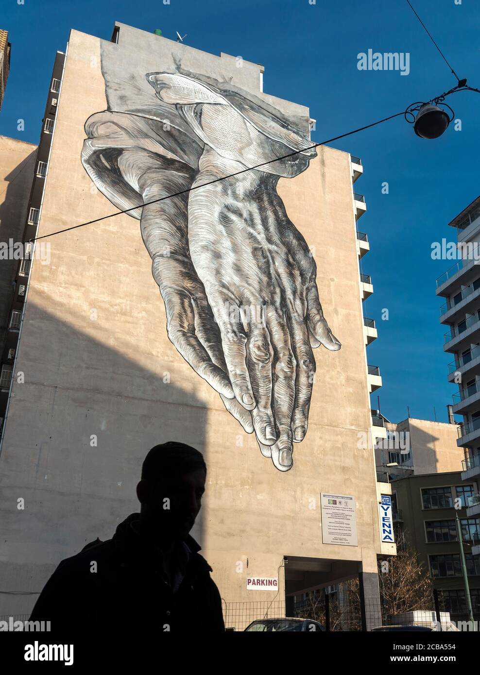 Über einem Passanten in der Pireos-Straße im Omonia-Viertel von Athen, Griechenland, ragt ein Street Art Wandbild in Form eines riesigen Paares betender Hände Stockfoto