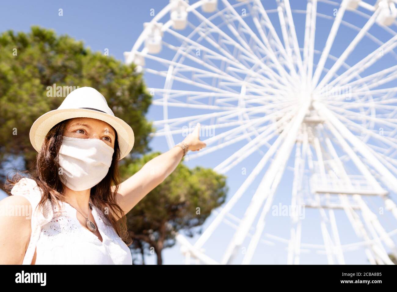 Kaukasische junge Frau mit weißem Hut und COVID Maske Schutz Zeigen auf das Riesenrad - Neue Tourismusregeln - Sommerurlaub mit Coronavirus saf Stockfoto