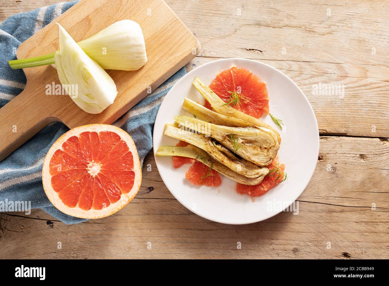 Sautiertes Fenchelgemüse mit Grapefruitscheiben, vegetarische Vorspeise auf weißem Teller, Zutaten und ein blaues Küchentuch auf rustikalem Holz, Kopie spc Stockfoto