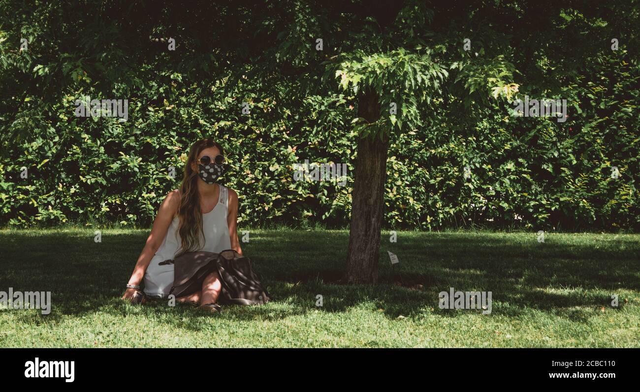 Horizontale Fotografie der jungen Frau in weißem Kleid, mit Sonnenbrille und Stoff Gesichtsmaske sitzen im Park unter einem Baum. Stockfoto