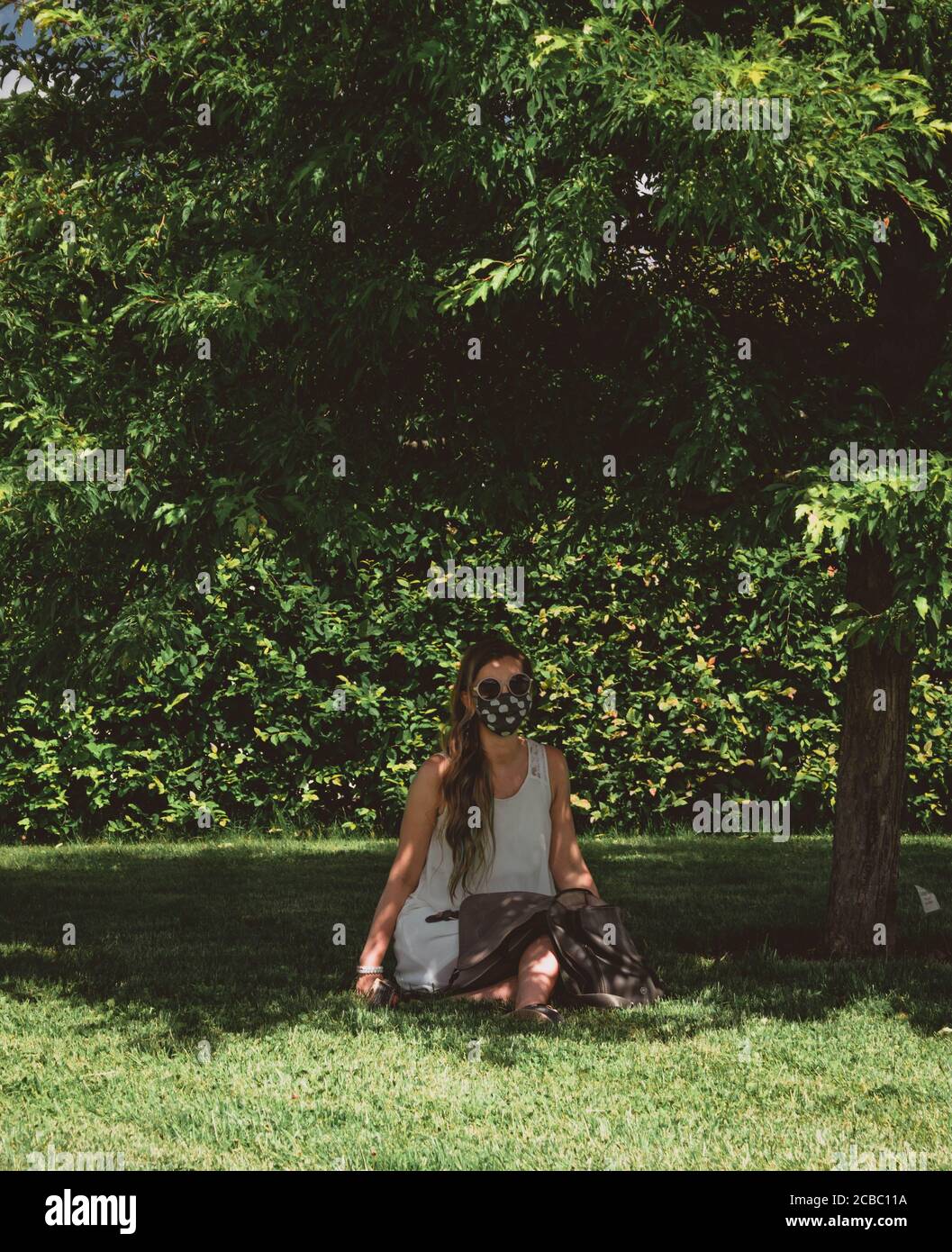 Vertikale Fotografie der jungen Frau in weißem Kleid, mit Sonnenbrille und Stoff Gesichtsmaske sitzen im Park unter einem Baum. Stockfoto