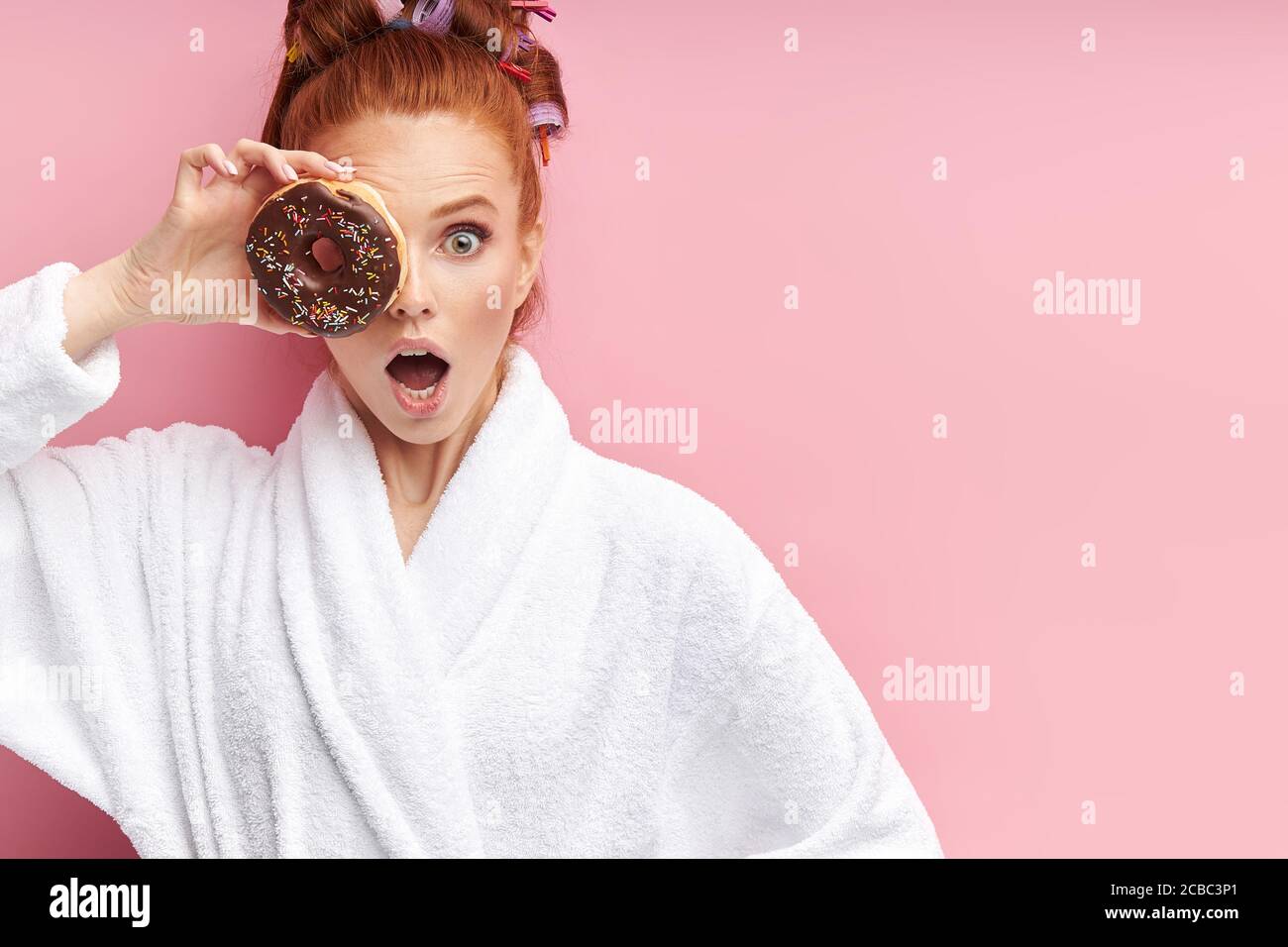 Mädchen nach der Dusche in Bademantel angezogen ist überrascht, geschlossen  rechtes Auge mit süßen braunen Donut. Isoliert auf rosa Hintergrund  Stockfotografie - Alamy