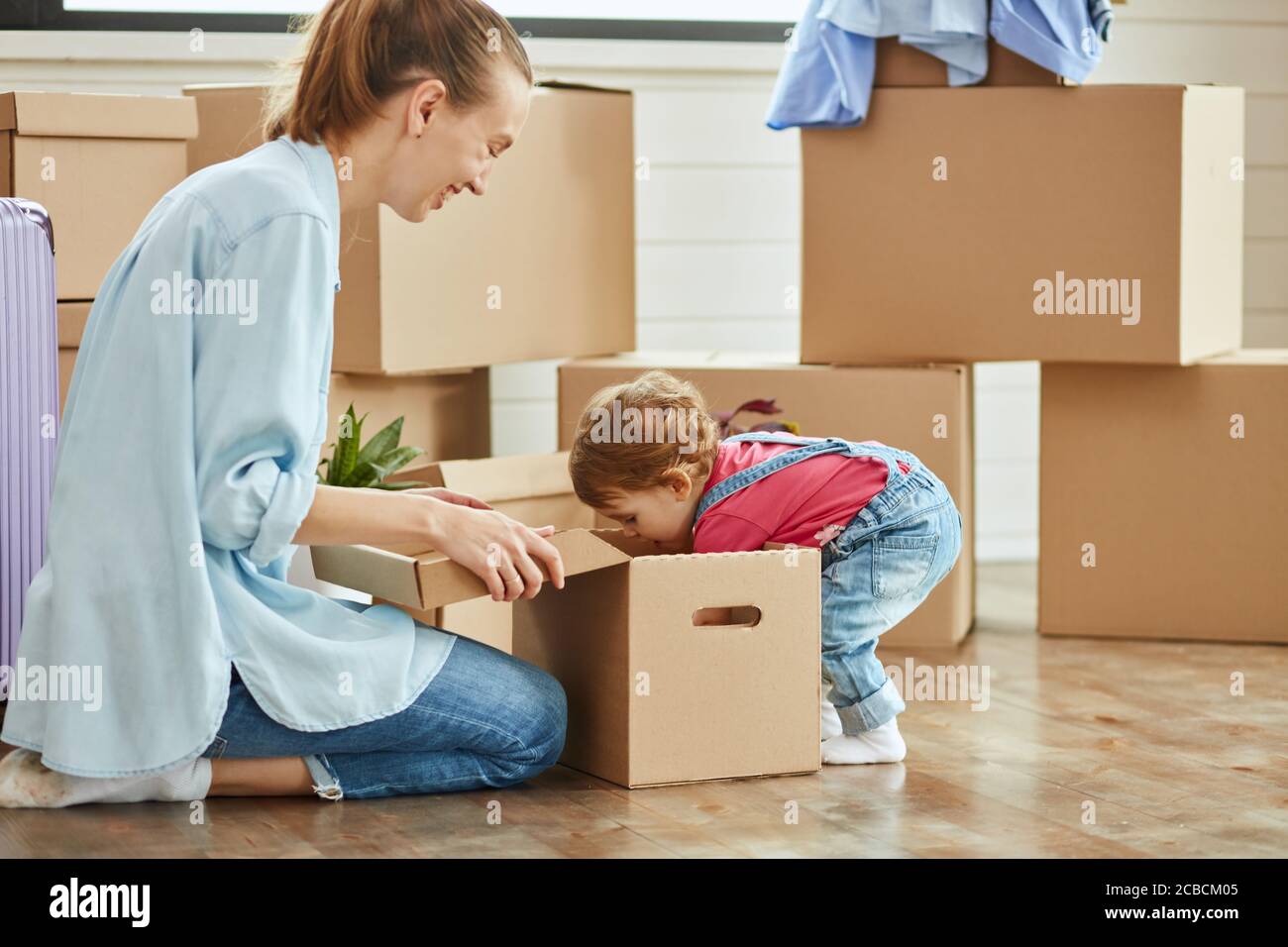 Kid tragen Denim Overall und blonde Frau tragen blaues Hemd, Jeans und weiße Socken Lächeln und spielen mit Box. Bewegliche Felder im Hintergrund Stockfoto