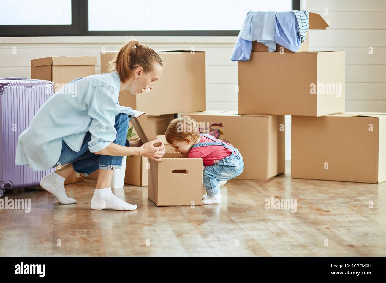In der Mitte Foto kleines Kind sieht in der Box. Mutter blonde Frau tragen blaues Hemd, Jeans und weiße Socken Lächeln und schauen Tochter. Hintergrund bewegt bo Stockfoto