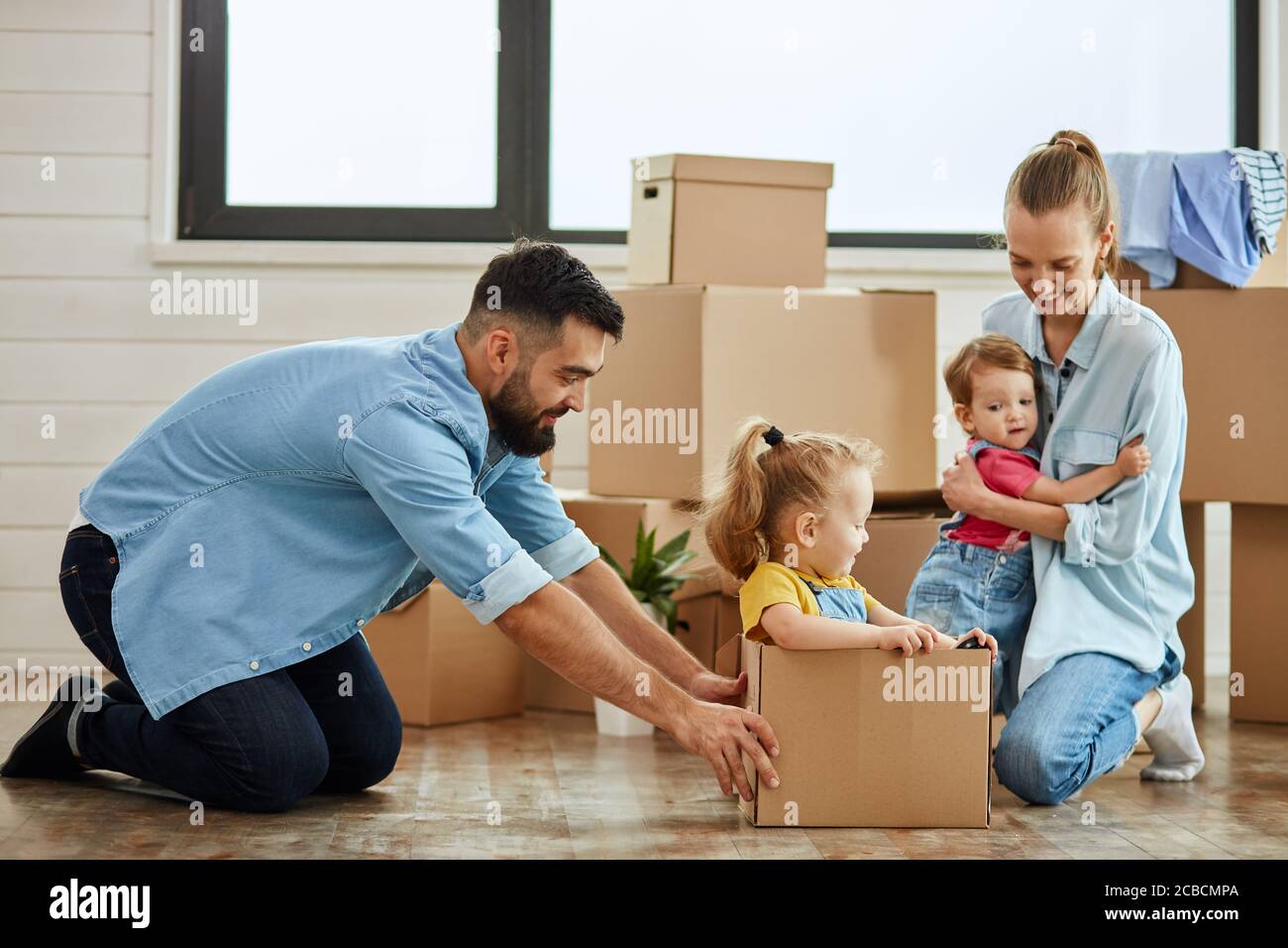 Kaukasische Familie, Mann, Frau und zwei Mädchen spielen mit Moving Box. Mutter umarmt jüngste Tochter, Vater saß jüngste Tochter in Box und bewegen Stockfoto