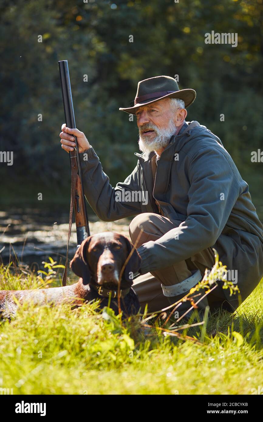 Senior gut aussehende Jäger mit seinem Tracker Hund auf der Jagd auf wilde Enten. In der Nähe des Sees sitzend, Hut tragend, Pistole haltend Stockfoto