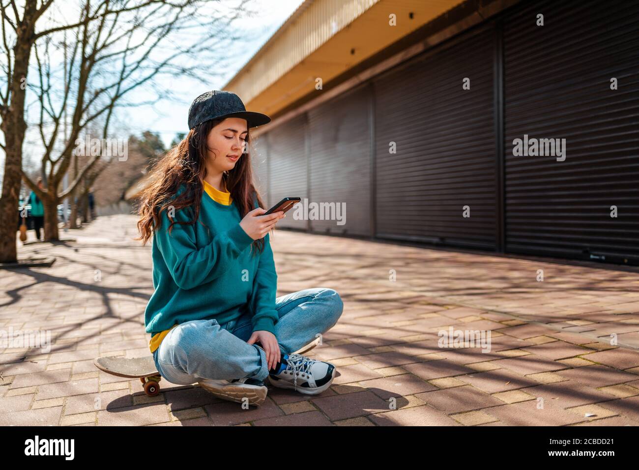 Eine junge Frau in legerer Kleidung sitzt mit gekreuzten Beinen auf einem Skateboard und nutzt ein Smartphone. Straße im Hintergrund. Technologie- und Kommunikationskonz Stockfoto