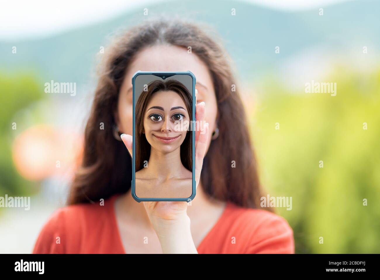 Eine Frau bedeckt ihr Gesicht mit einem Smartphone mit einem Cartoon-Porträt einer Frau. Das Konzept, Identität und gefälschte Seiten in sozialen Netzwerken zu verbergen. Stockfoto