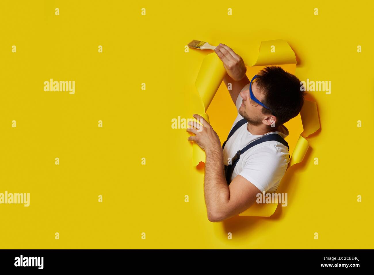 kaukasischen Maler Arbeiter in Sicherheitsglas und Uniform halten Pinsel durch Loch in gelbem Papier. Handwerker Malerei Wand. Kopieren Raum Stockfoto