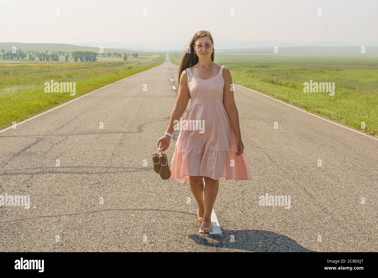 Eine weiße Frau in einem rosafarbenen Sundress mit lockeren Haaren läuft barfuß mitten auf einer Asphaltstraße und hält Sandalen in den Händen. Stockfoto