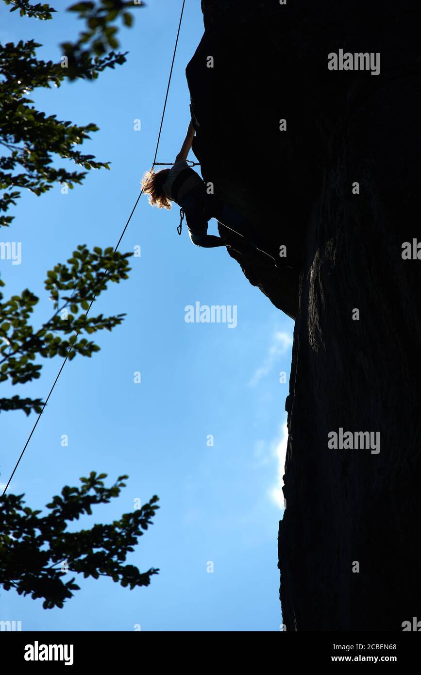 Vertikale Momentaufnahme einer Silhouette weibliche Figur, die den dunklen Felsen hinauf klettert, die Spitze erreicht, gegen blauen Himmel, grüne Blätter unten. Low-Angle-Ansicht. Konzept des Bergsports, Bergsteigen, Alpinismus. Stockfoto