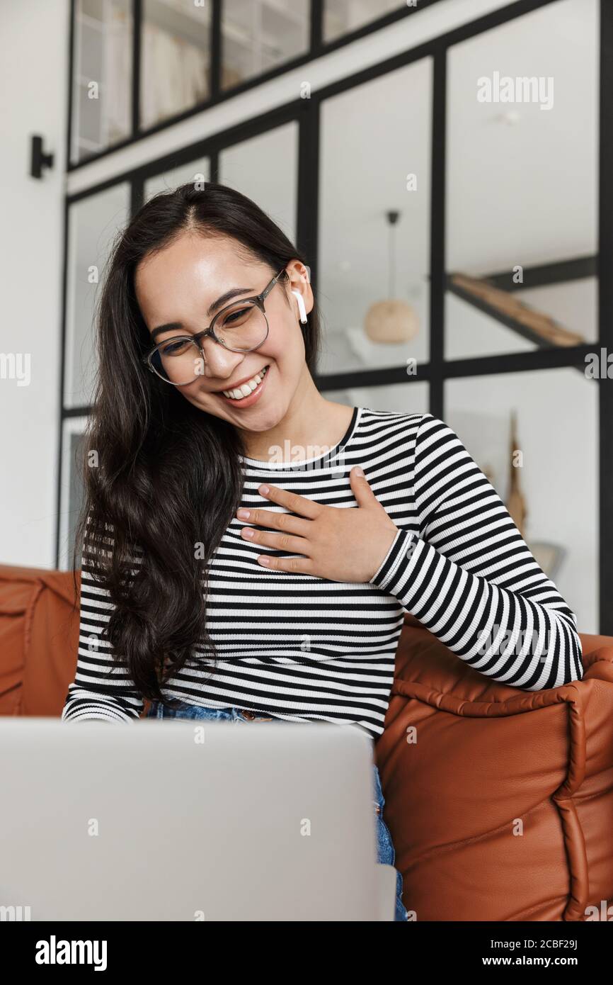 Attraktive lächelnde junge asiatische Geschäftsfrau entspannen auf einer Ledercouch zu Hause, während Videoanruf auf Laptop-Computer, tragen Kopfhörer Stockfoto