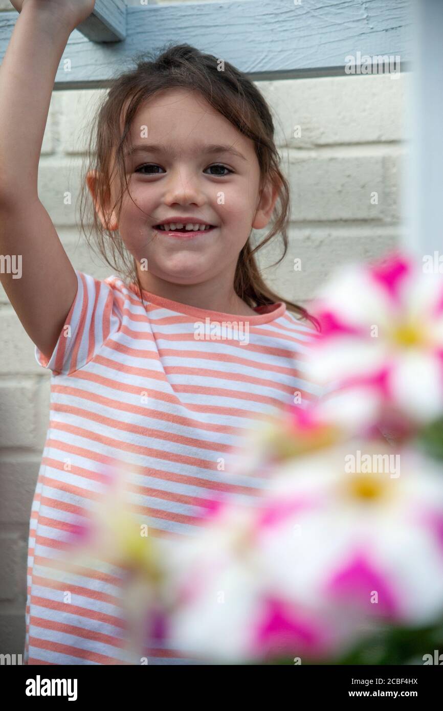 4 Jahre altes weißes Mädchen, in gestreiftem T-Shirt und Armen über dem Kopf, spielt in einem Garten voller rosa und weiß gestreifter Petunia Blumen. Lancashire, England Stockfoto