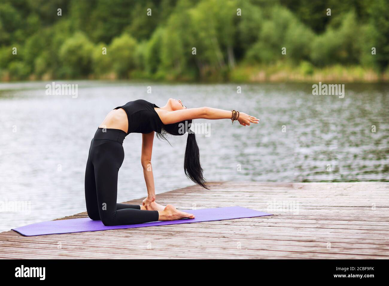 Brunette Mädchen mit langen Haaren in einem schwarzen Sportanzug macht Übungen, sitzt auf einer Matte auf Holzbrettern am Fluss ausgelegt Stockfoto