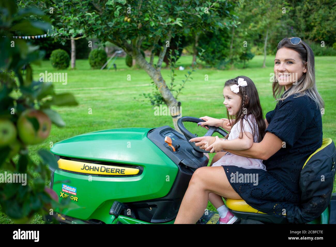 Mutter und junge Tochter, die einen grünen und gelben John Deere Traktormäher in einem großen englischen Obstgarten fahren. Lancashire, England, Großbritannien Stockfoto