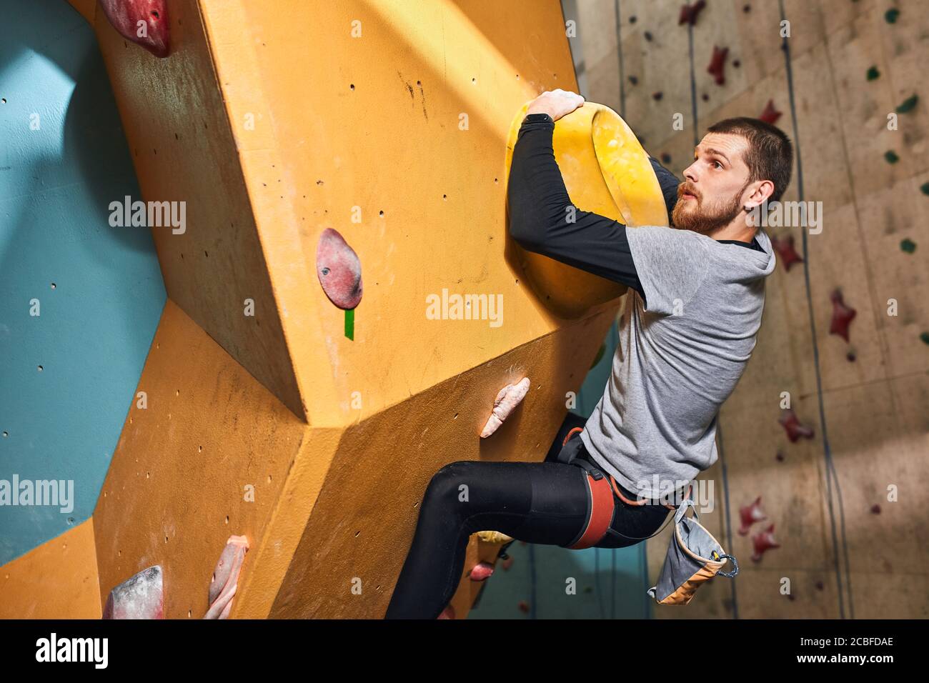 Seitenansicht Aufnahme von jungen schönen Boulderer mit Bart, hängen auf großen Halt an bunten Kletterwand, Denken an seinen nächsten Schritt, wählte kompliziert Stockfoto