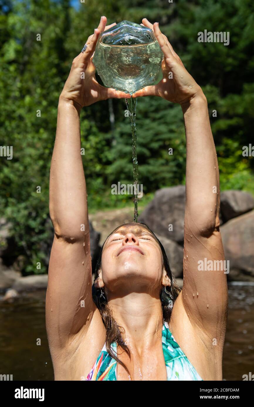 Nahaufnahme einer jungen Frau mit den Armen nach oben, die Wasser aus einem Glasbehälter in einem Bergbach oder Fluss auf den Kopf und das Gesicht gießt. Stockfoto