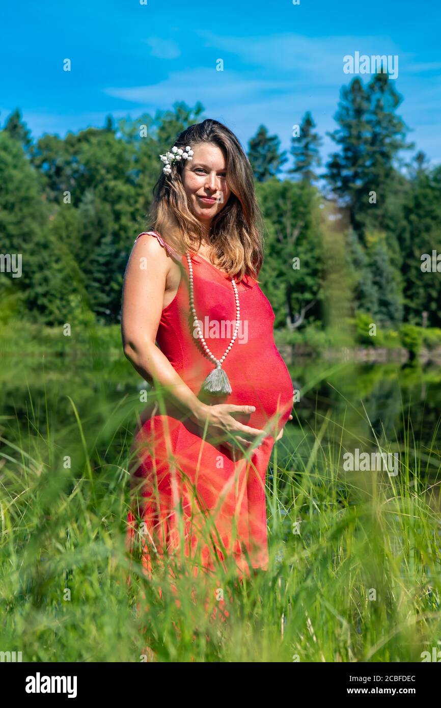 Junge schwangere Frau in einem ärmellosen roten Kleid, die am Ufer eines kanadischen Sees steht, zärtlich ihren Bauch hält und die Kamera anlächelt, eine Blume im Haar. Stockfoto