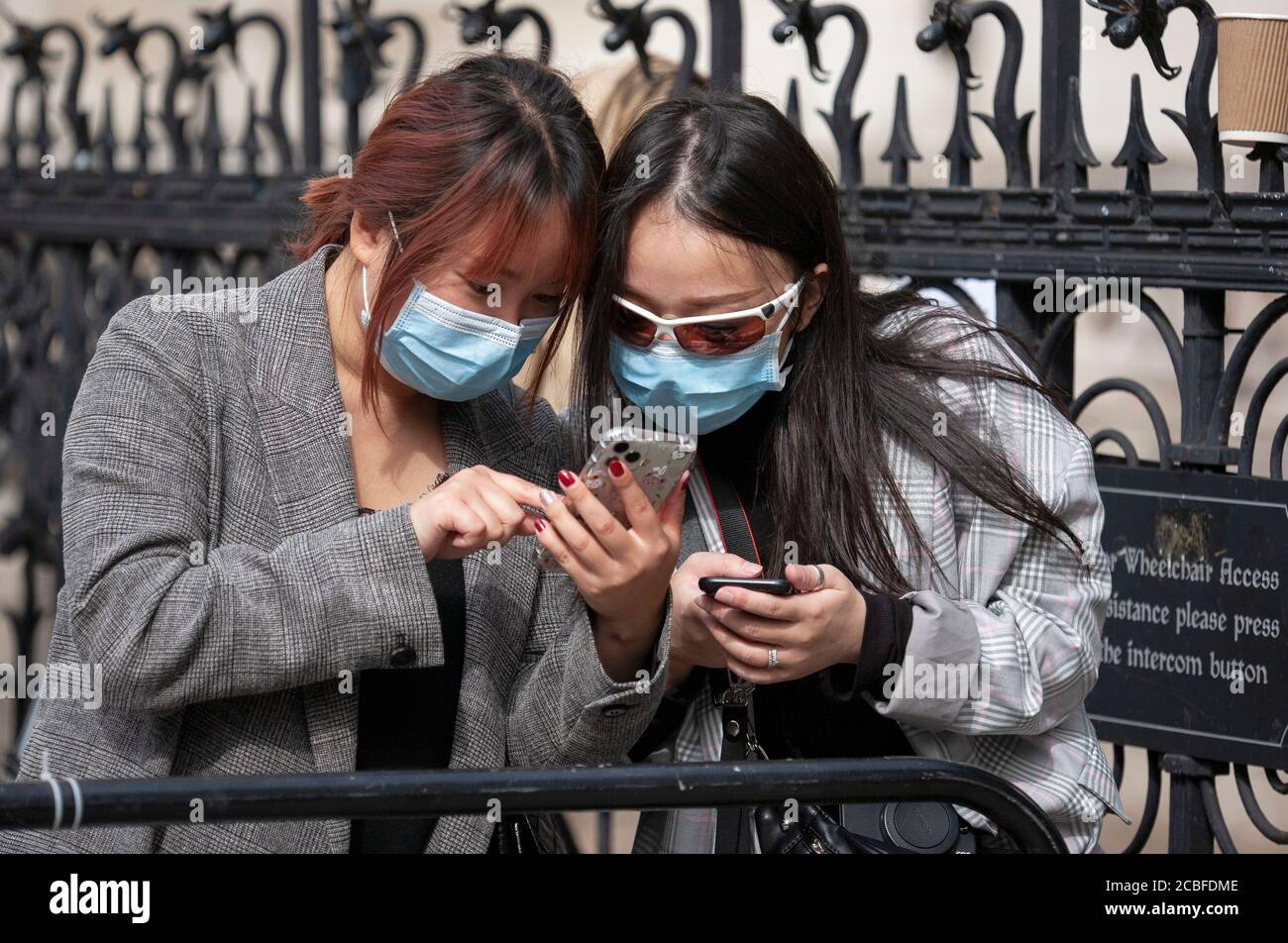 Zwei asiatische Frauen, die Gesichtsmasken tragen, schauen sich ihre Handys außerhalb des Hofes an. London, England Stockfoto