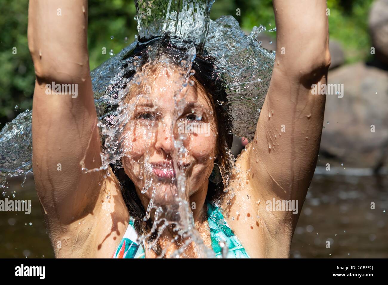 Nahaufnahme des Gesichts einer jungen Frau mit den Armen nach oben, die aus einem Glasbehälter in einem Bergfluss Wasser auf sich selbst gießt und die Kamera anlächelt Stockfoto