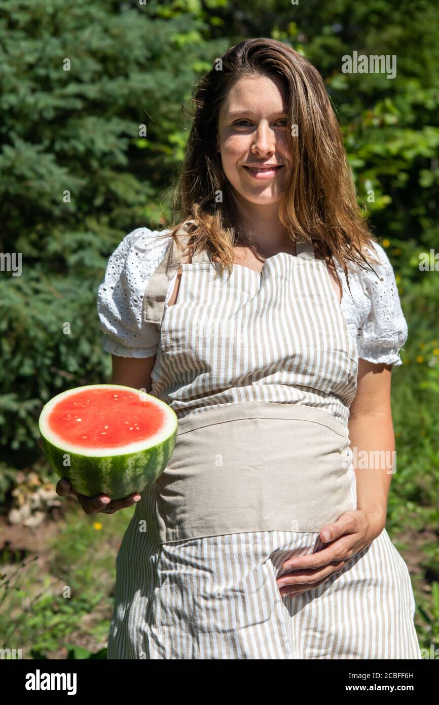 Junge, schöne schwangere Frau posiert mit einer halben, saftigen und roten Wassermelone neben ihrem großen Bauch. Waldhintergrund, nicht urbanes Porträt. Stockfoto