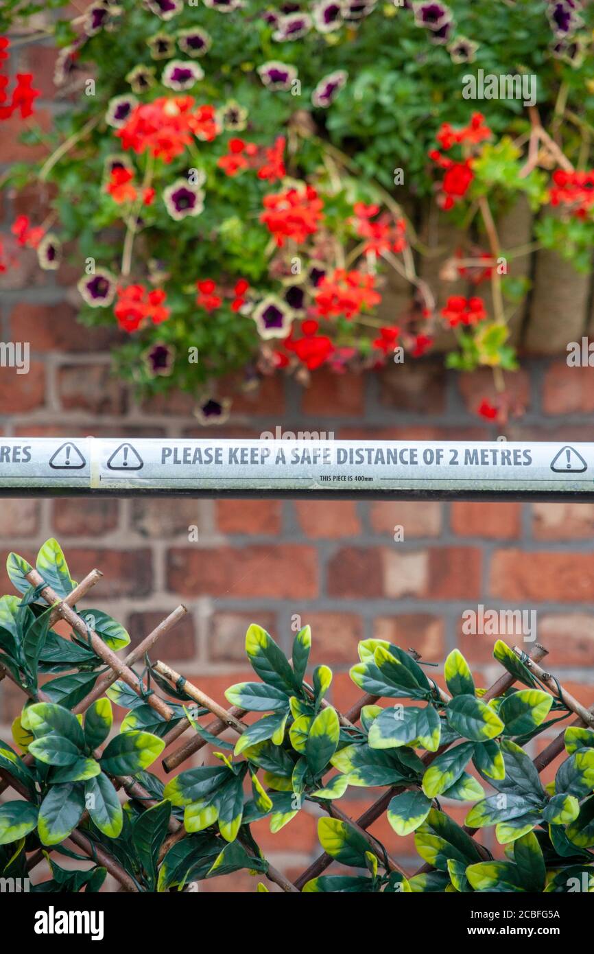 ‘Bitte halten Sie einen Sicherheitsabstand von 2 Metern’ Warnband vor einer Ziegelwand mit hängendem Korb mit Sommerblumen. England GB Stockfoto