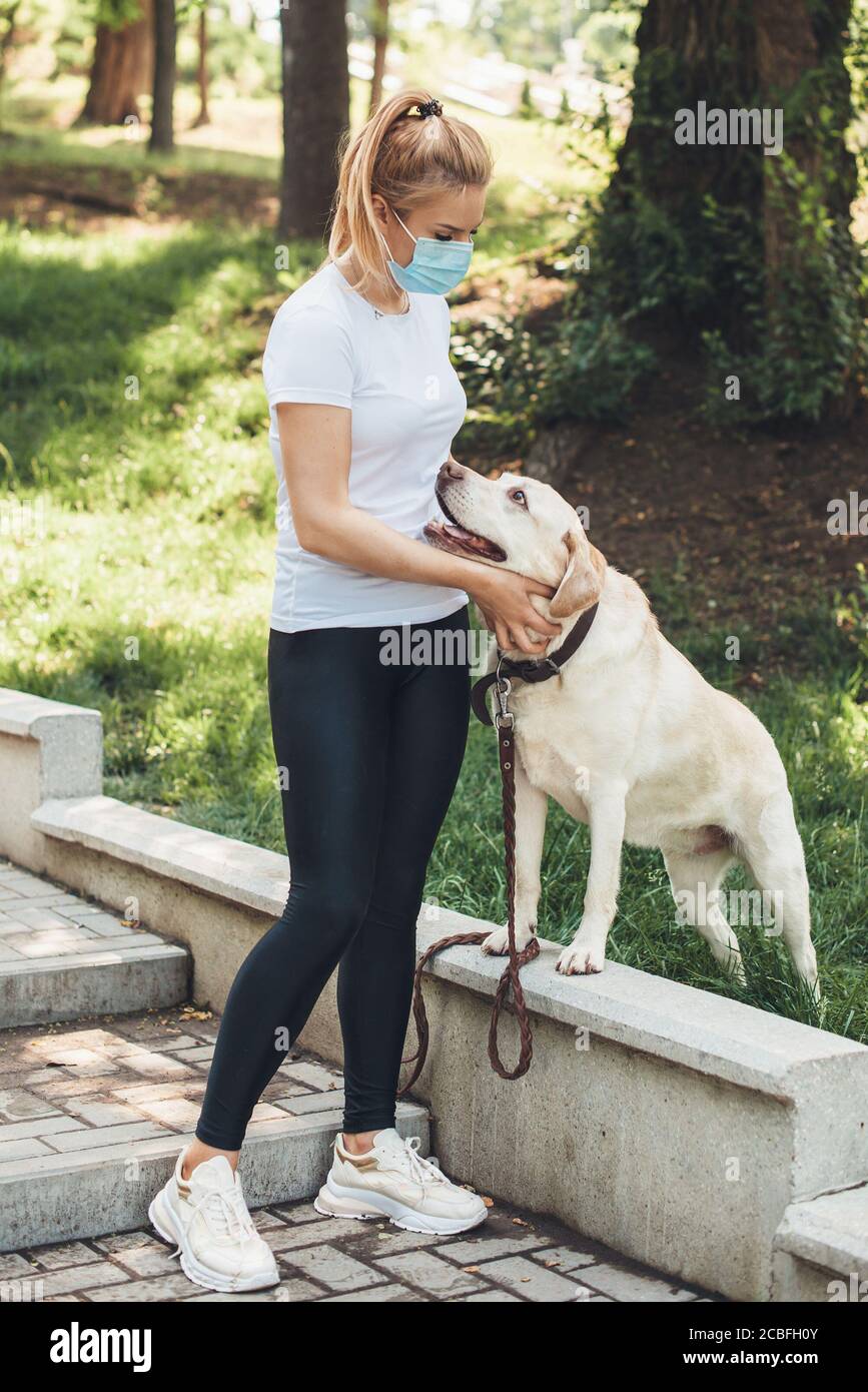 Kaukasische blonde Dame mit medizinischer Maske auf Gesicht zu Fuß mit Ihr Hund draußen im Park Stockfoto