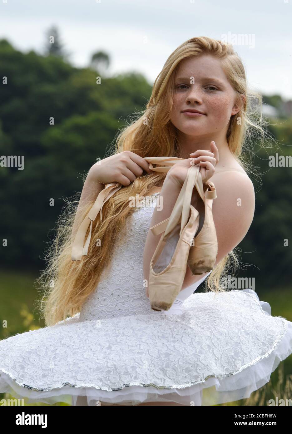 Eine Ballerina mit langen blonden Haaren und Sommersprossen starrt in die Kamera und hält ihre spitzenschuhe. Sie trägt ein weißes Tutu. Stockfoto