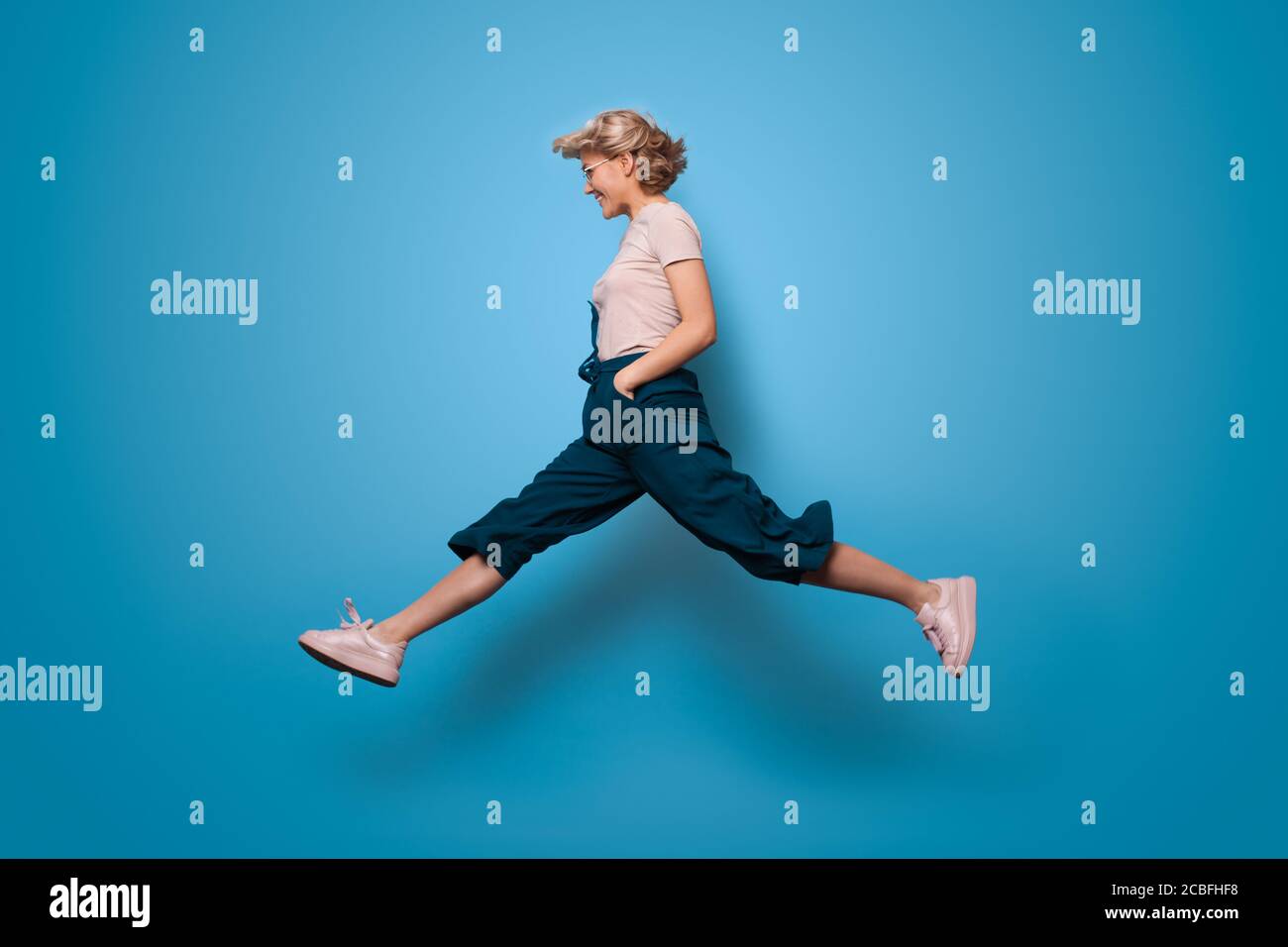Charmante blonde Dame springt auf einem blauen Studio Wand und Lächeln Sie Werbung für etwas Stockfoto