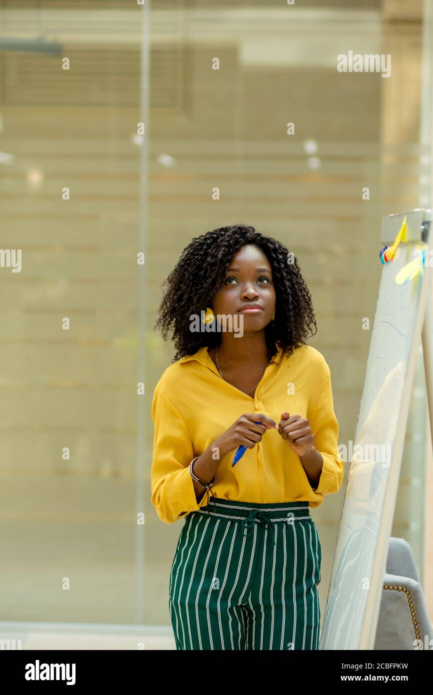 Schöne nachdenkliche afroamerikanische Frau mit einer wilden Afro-Frisur, die neben dem Flipchart steht und nach oben schaut Stockfoto