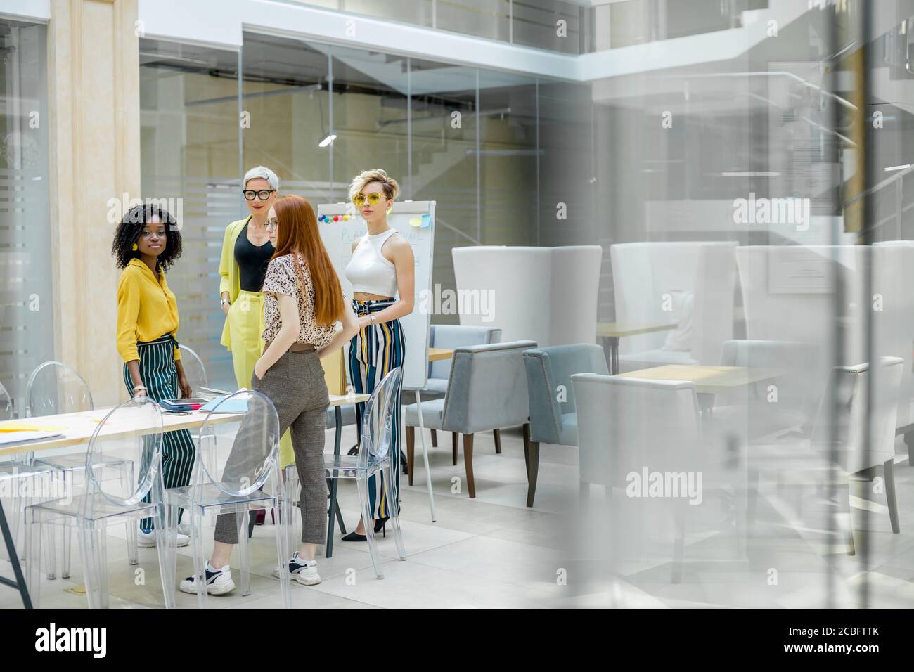 Multiethnische Gruppe von lächelnden jungen Frauen stehen, reden, warten auf Chef im Konferenzraum. Kopierraum. Glaswand Stockfoto