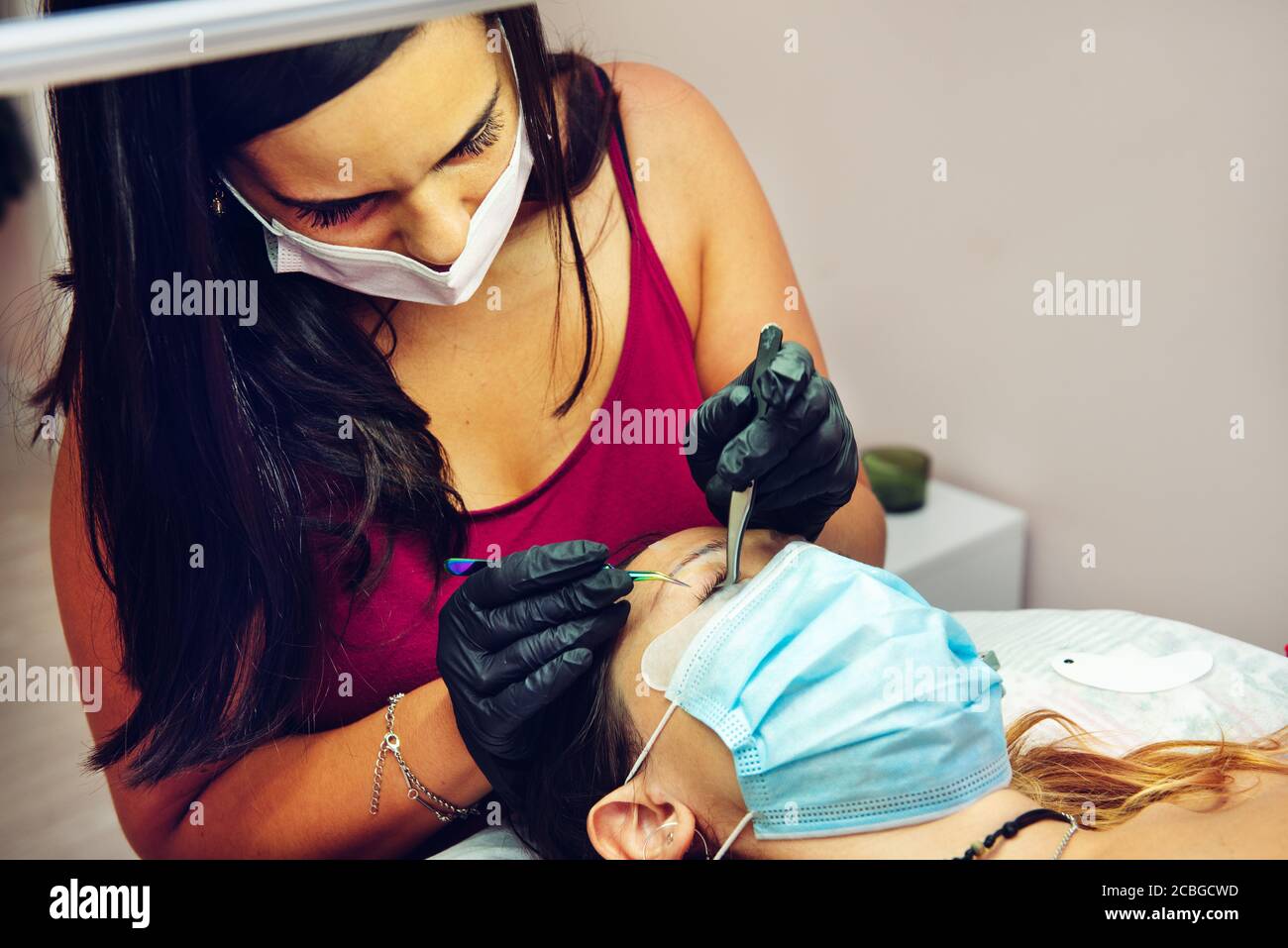 Junge langhaarige Brünette Frau setzen falsche Wimpern auf Dame im Schönheitssalon mit Masken, neue normale Stockfoto