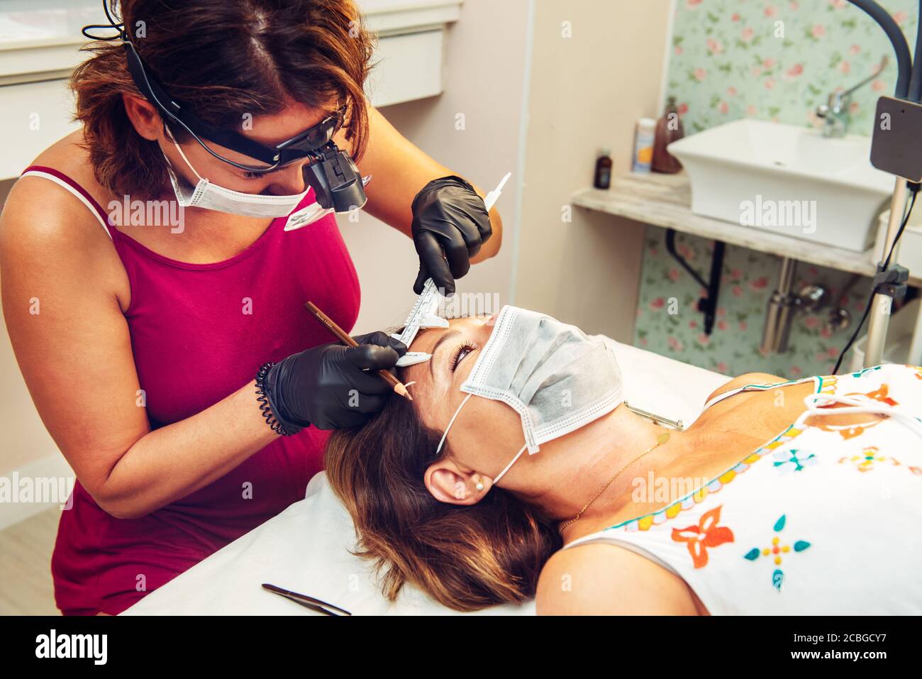 Kosmetikerin im Beauty-Center, die Messungen an Kunden für eine Augenbraue Behandlung, mit Masken während der neuen Normalität Stockfoto