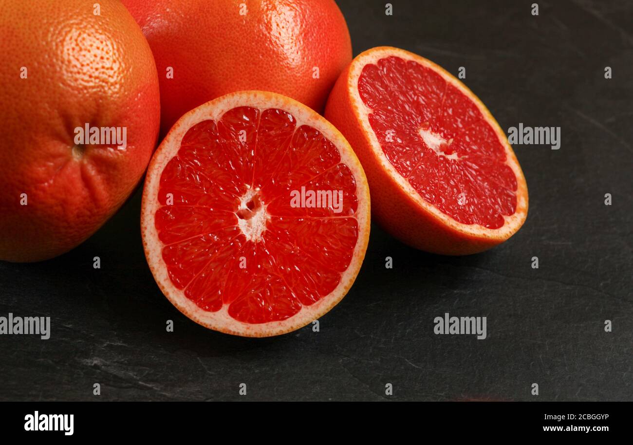 Rosa Grapefruit zur Hälfte geschnitten, mehr Zitrusfrüchte im Hintergrund, auf schwarzem Brett. Stockfoto