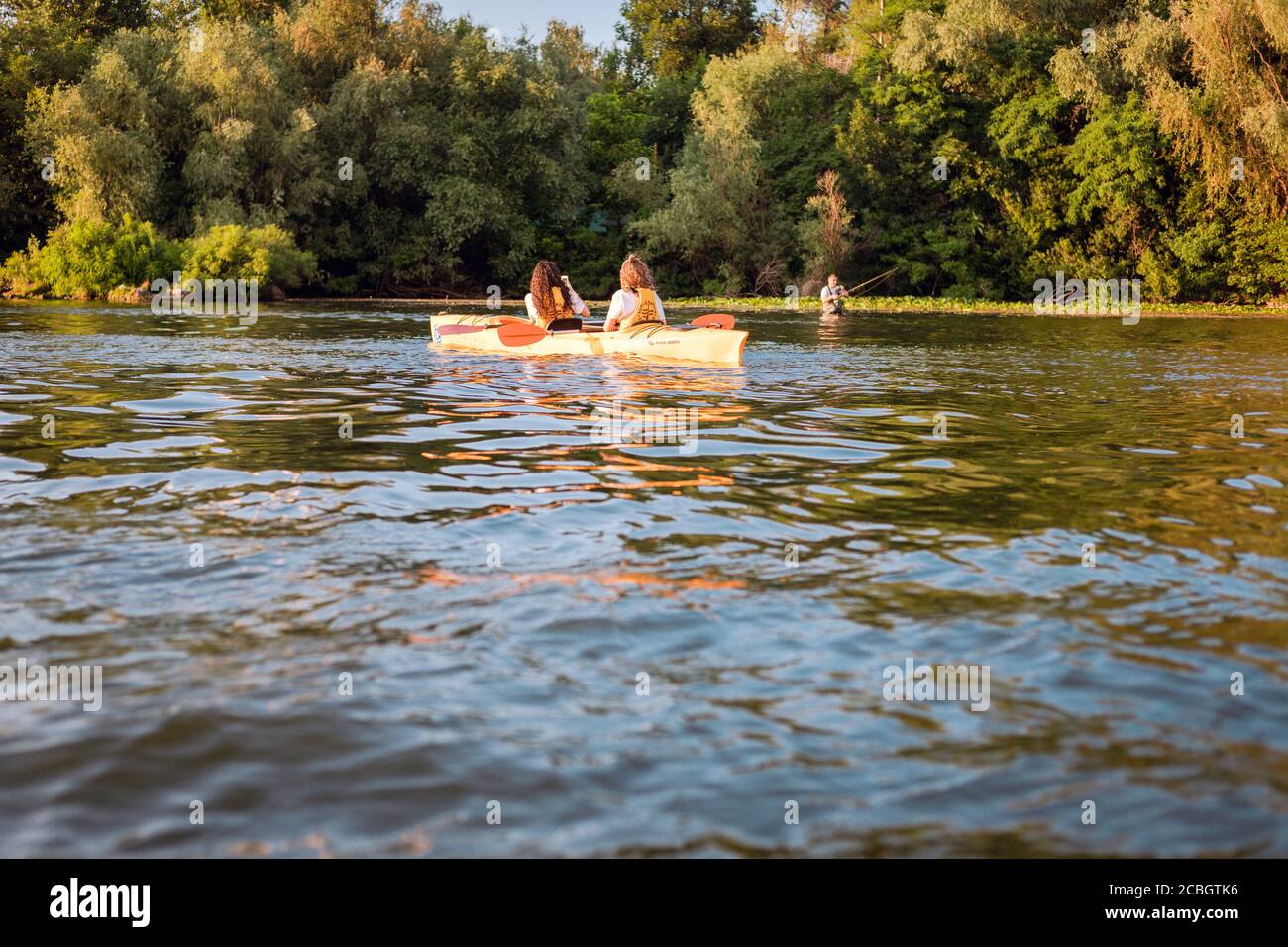 Zwei Mädchen fotografieren sitzen im Kajak auf dem Fluss auf dem Hintergrund des Mannes Angeln in der Nähe der Küste bei Sonnenuntergang in sonnigen Sommertag. Hobby-Konzept, Sport Stockfoto