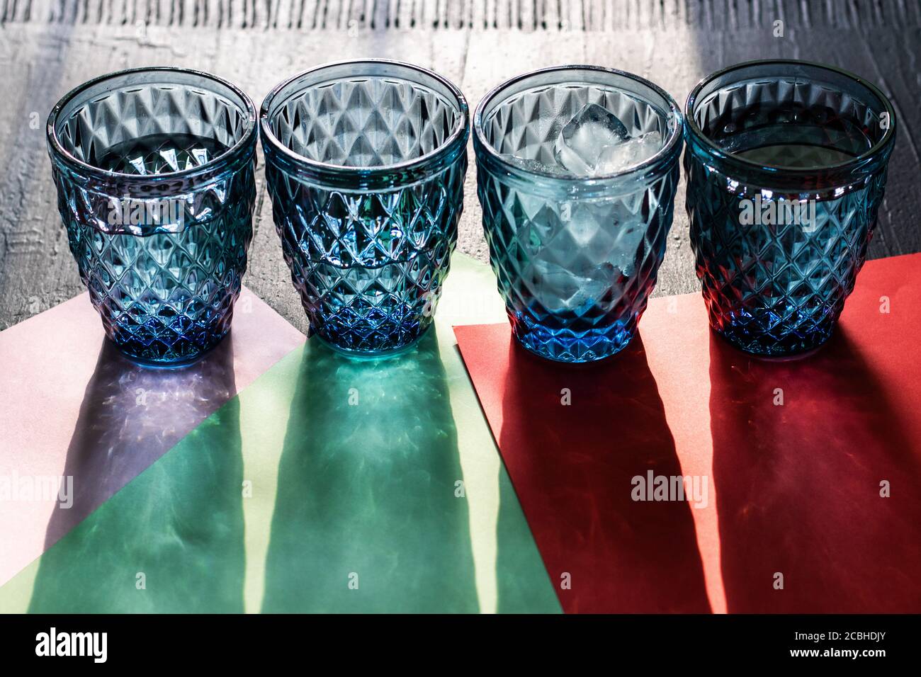 Vier gemusterte blaue Gläser Wasser mit Eis oder Cocktails mit langen Schatten auf farbigem Papier und schwarzem Hintergrund. Stockfoto