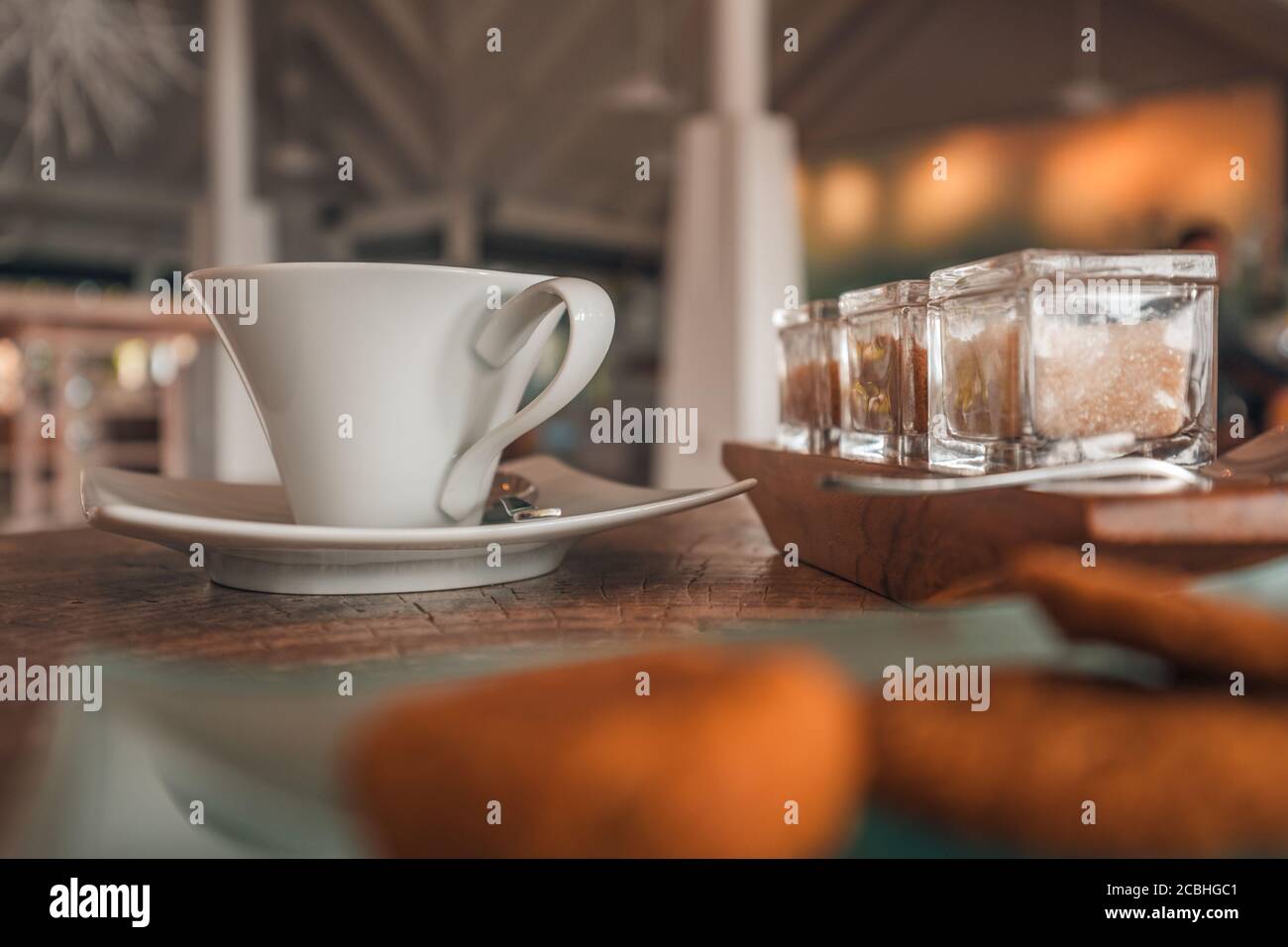 Niedriger Blick auf Holztisch mit weißem Kaffeebecher, braunem Zucker und Keksen. Entspannender Coffee Shop in der Nähe, Morgenlicht, positive Vibes Stockfoto