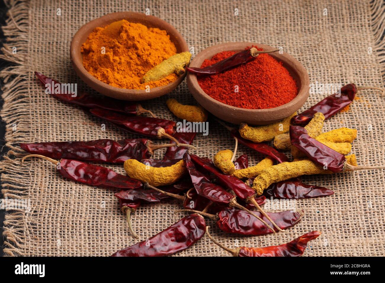 Indische Gewürze Chili und Kurkuma Pulver in einem Tontopf mit roten Chilis und Kurkuma Wurzel auf Jute Hintergrund. Stockfoto
