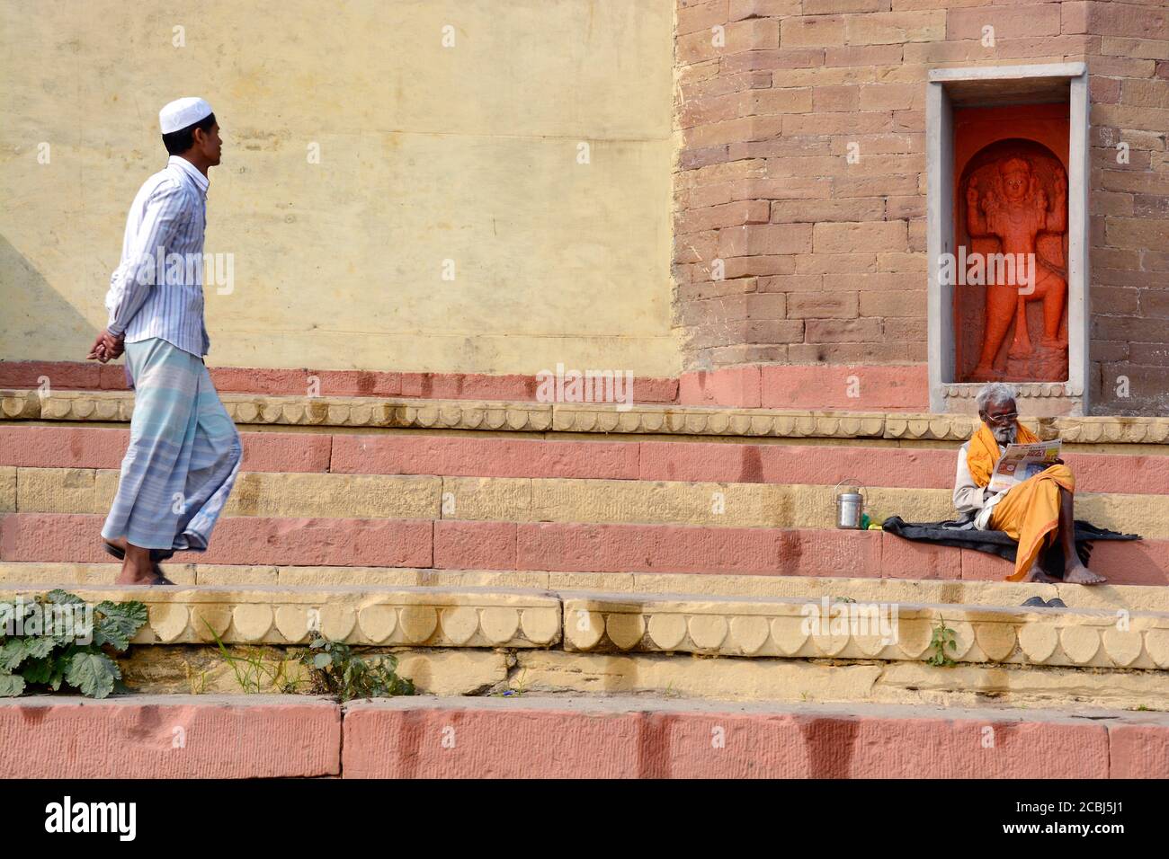Varanasi Ganga Ghat EIN Hindu-heiliger sitzt vor dem Hanuman-Tempel und ein muslimischer Junge geht durch das Ghat. Stockfoto