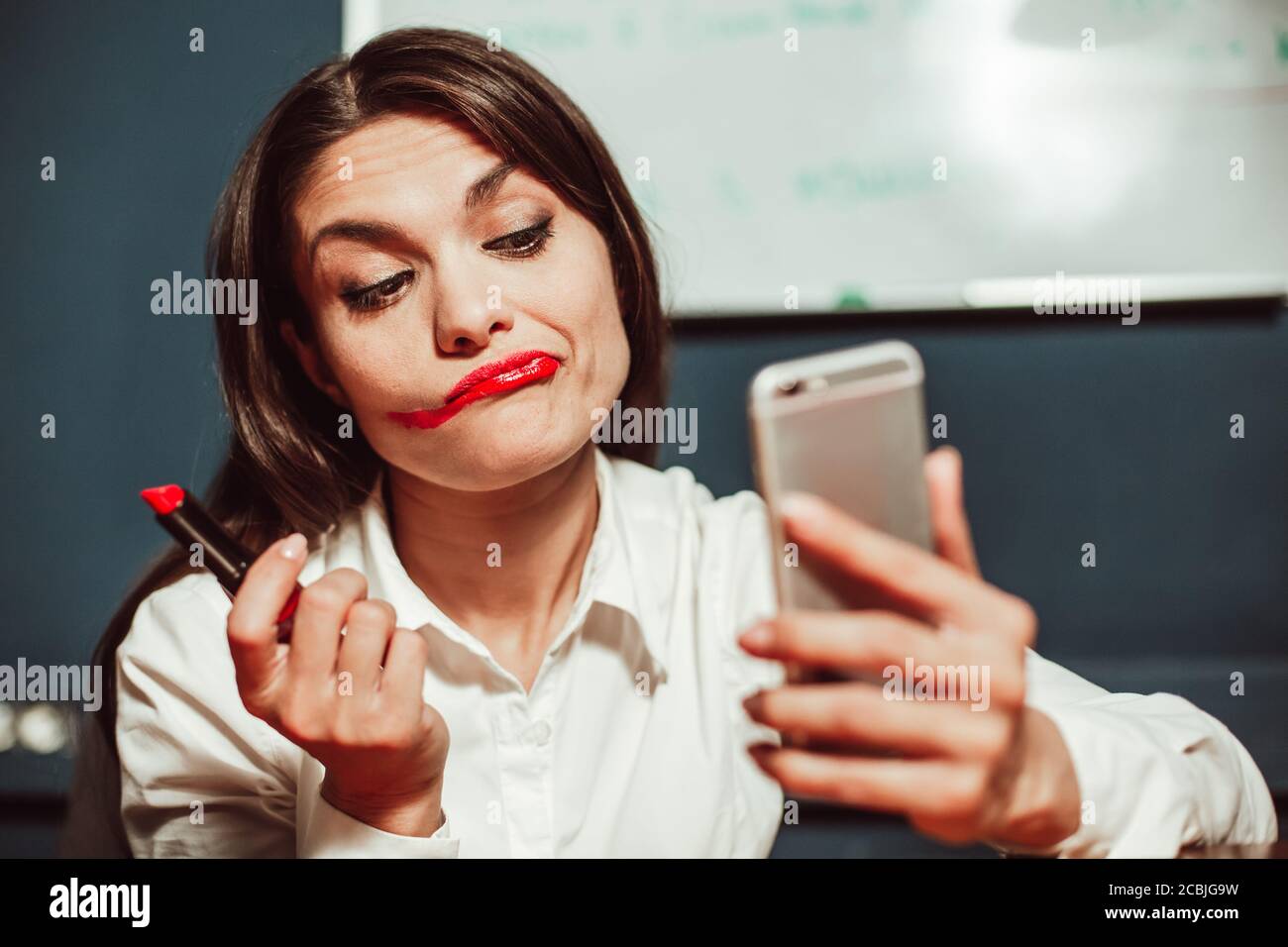 Verrückte Büroleiterin Geschäftsfrau macht Make-up mit roten Lippenstift am Arbeitsplatz mit Handy statt Spiegel. Getöntes Bild Stockfoto