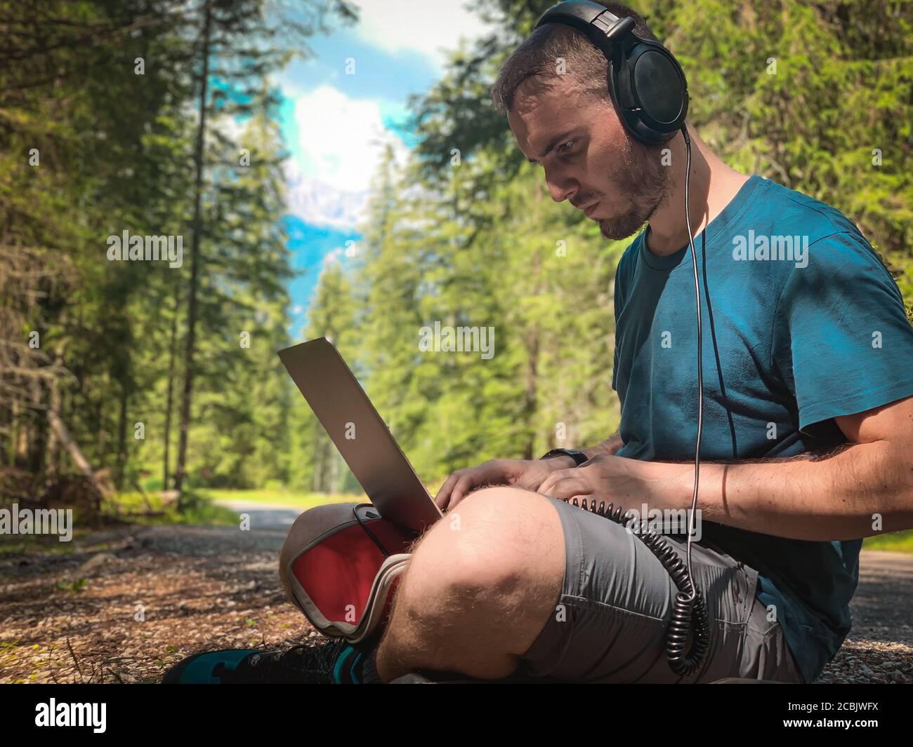Junge Brünette kaukasischen Mann mit Bart tragen Kopfhörer und die Arbeit auf Laptop in einem Berg Ort, grünen Wald im Hintergrund, Konzept der smart Stockfoto